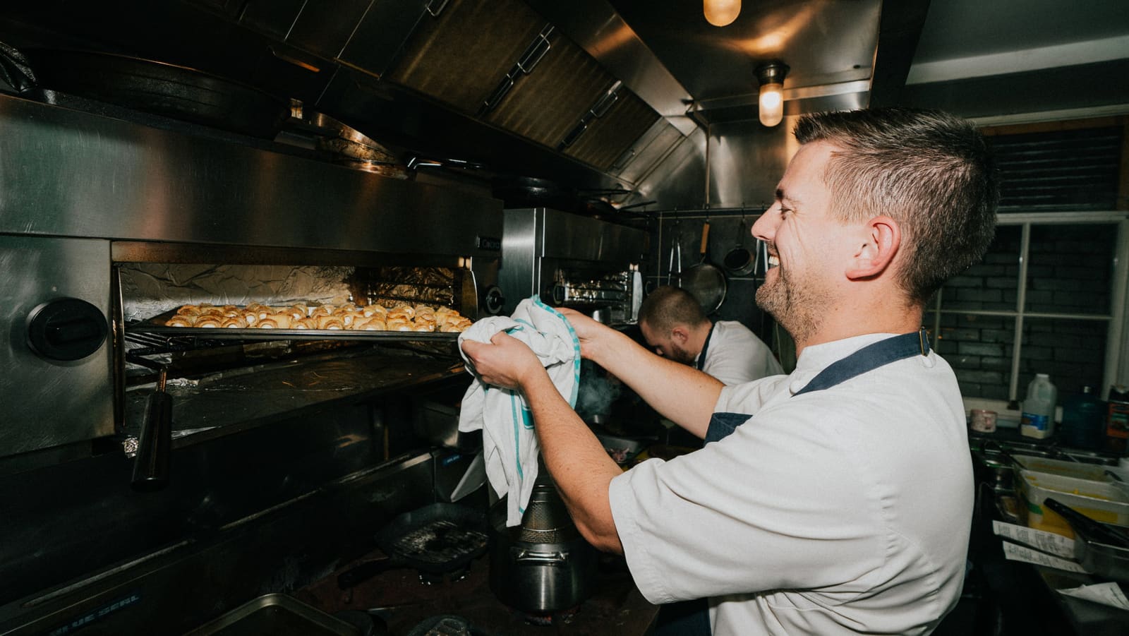 Chef Robert Thompson MBE smiling at the oven in the Royal Maritime House kitchen, a member of the brigade behind him